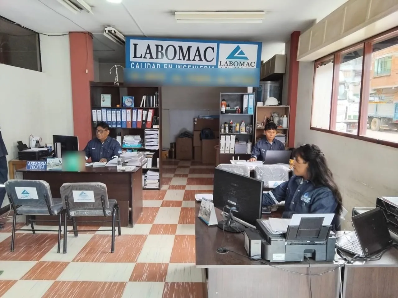 Oficina de LABOMAC con tres personas trabajando en sus escritorios, archivadores al fondo y el letrero de la empresa.